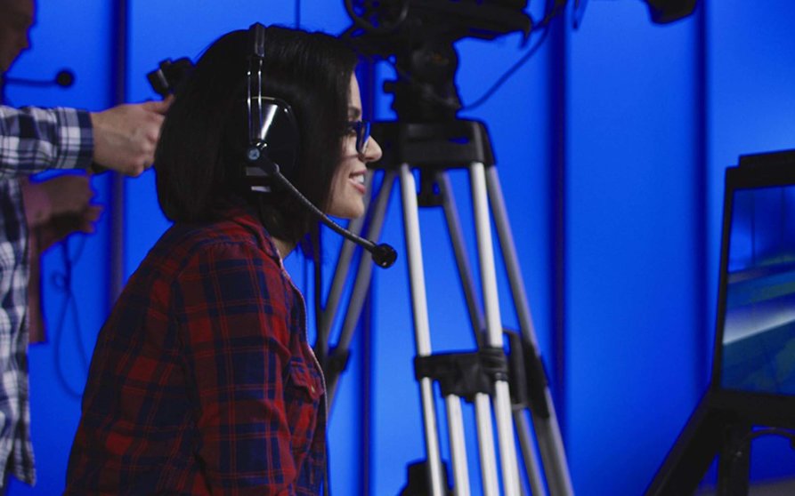 Woman wearing a headset sitting behind a monitor with the cameraman during production giving instructions in a newsroom gesturing and pointing