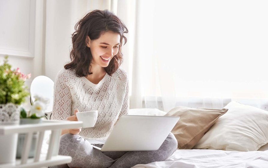 Happy casual beautiful woman working on a laptop sitting on the bed in the house.
