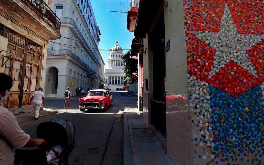 Un vehículo clásico y varios peatones pasan hoy por una calle con murales de la bandera cubana, junto al Capitolio en La Habana (Cuba). EFE/ Ernesto Mastrascusa
